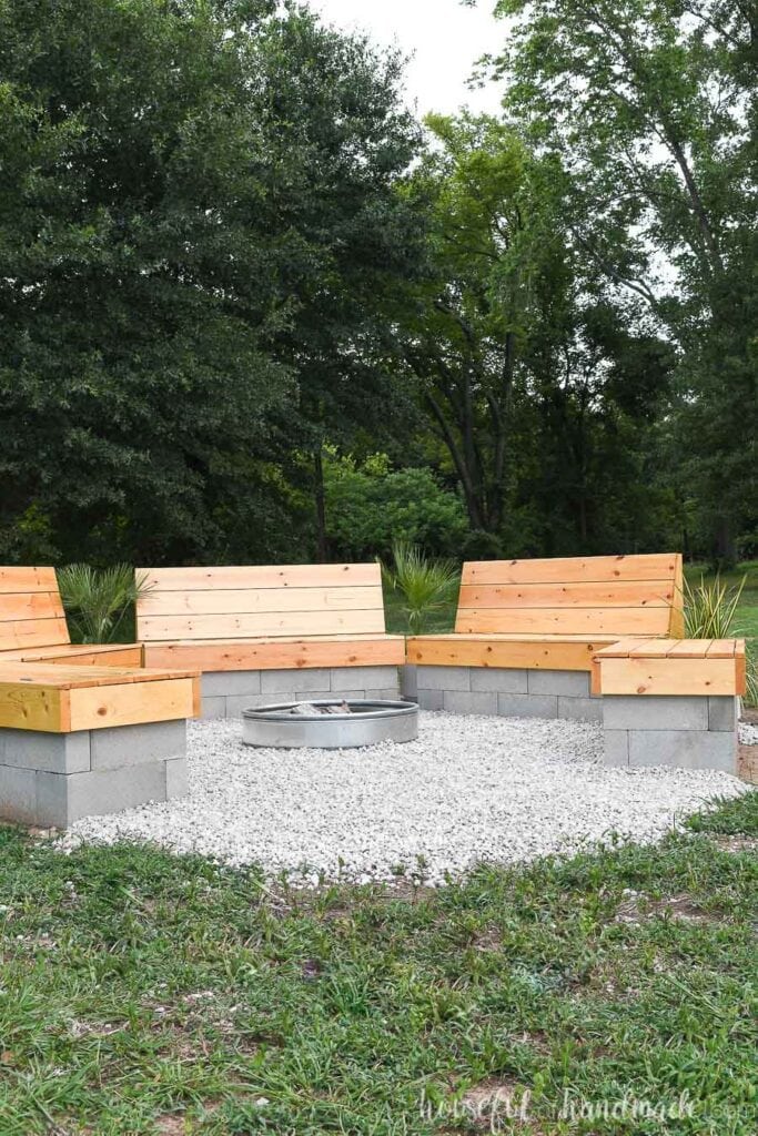 Five benches in a hexagon around a fire ring with gravel between them and wooded area in the background.