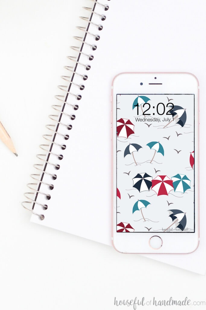 White iPhone with free Digital wallpaper for July on the screen sitting on a notebook.