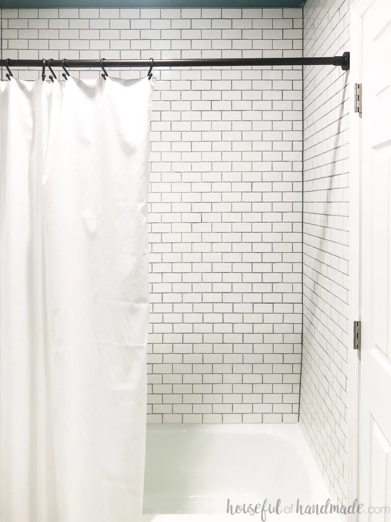 Shower tiled with small white ceramic subways tiles and rubbed bronze shower curtain rod with white shower curtain. 