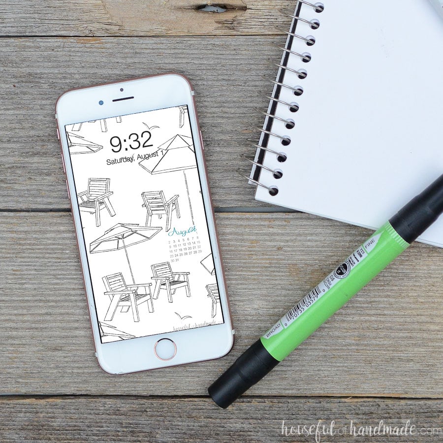 Smartphone on a reclaimed wood table with free digital wallpaper for August on the background next to a sketchbook.