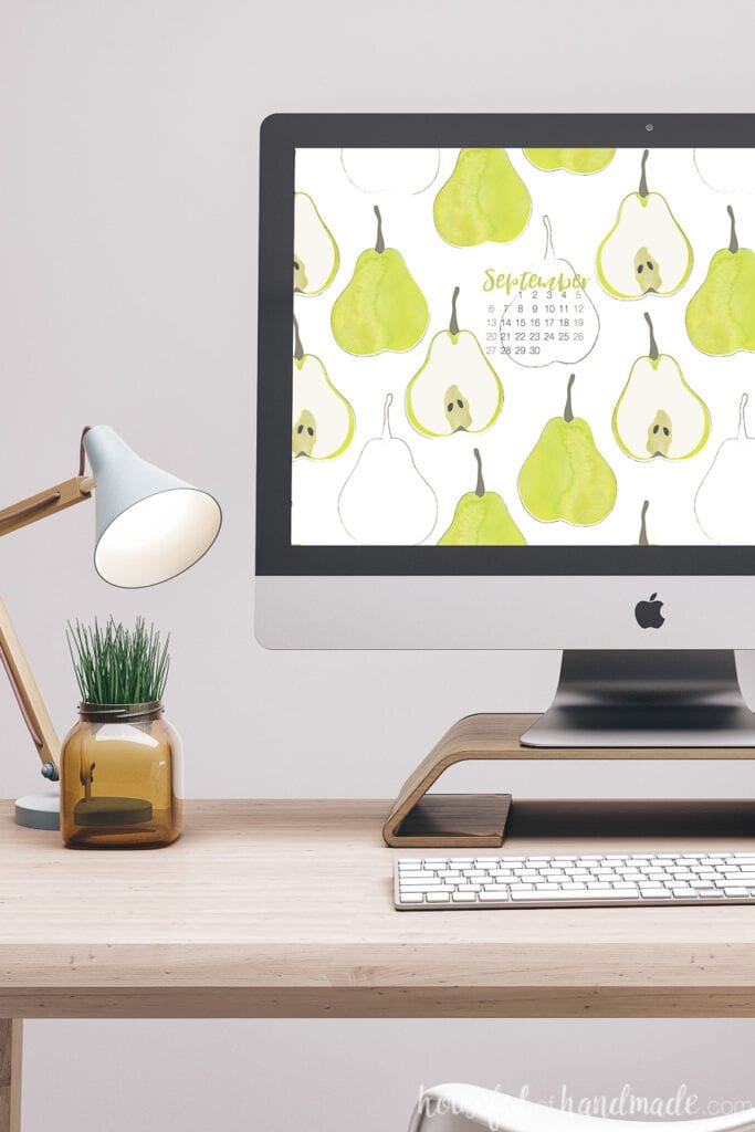 Desktop computer with the free digital wallpaper for September with a September calendar on the screen sitting on a desk with a desk lamp and plant.