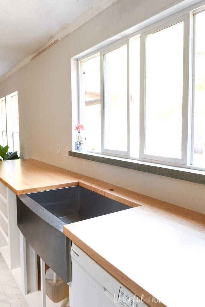 DIY wood countertops made from hard maple on top of white kitchen cabinets with a stainless steel farmhouse sink and large window in the wall above. 