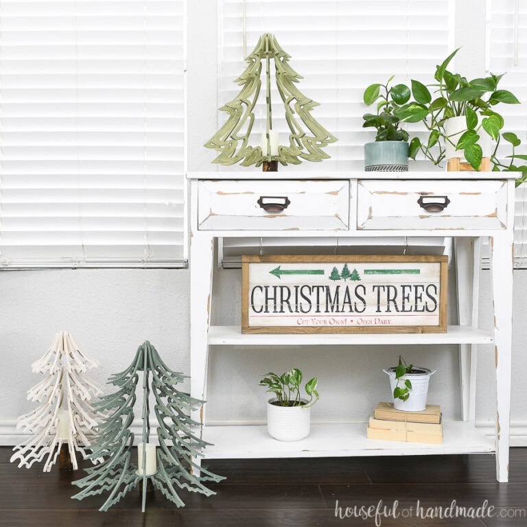 Three 3D wood Christmas tree lanterns sitting on and next to a small white console table.