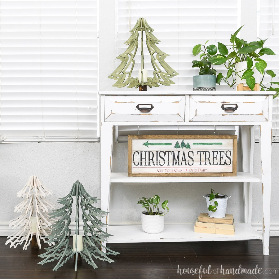 Three 3D wood Christmas tree lanterns sitting on and next to a small white console table. 