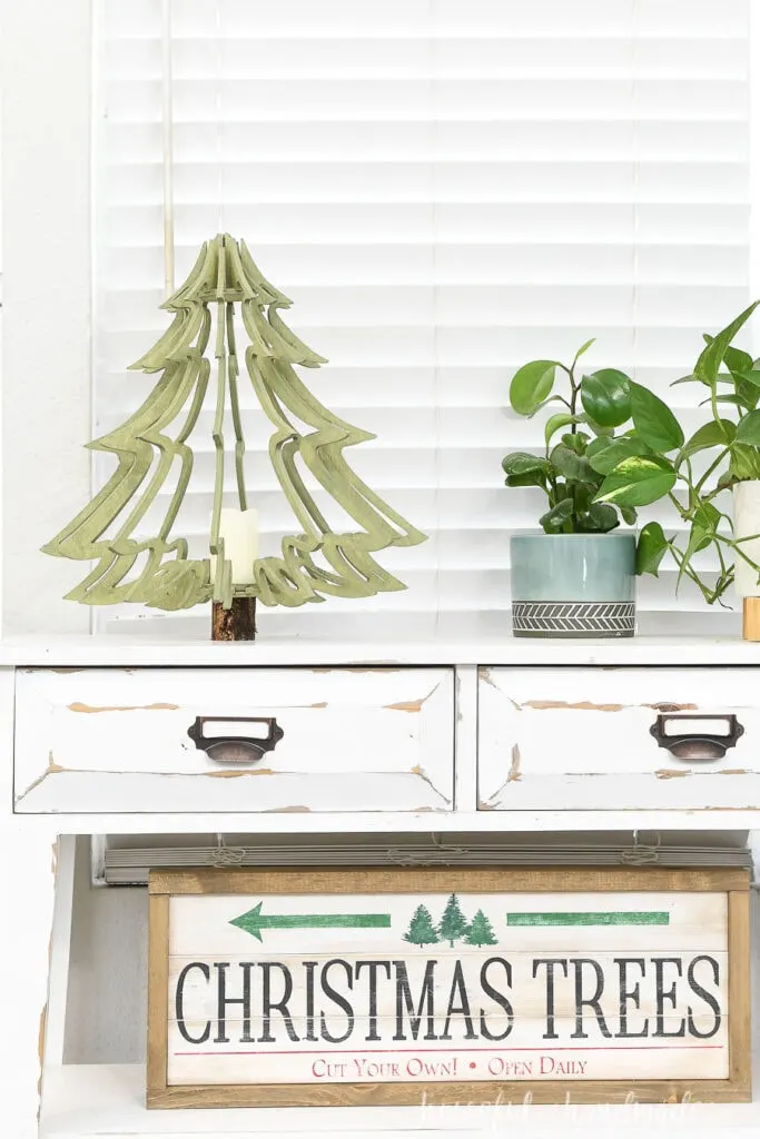 Large table top Christmas tree made from plywood on a console table next to house plants and above a Christmas tree wood sign. 