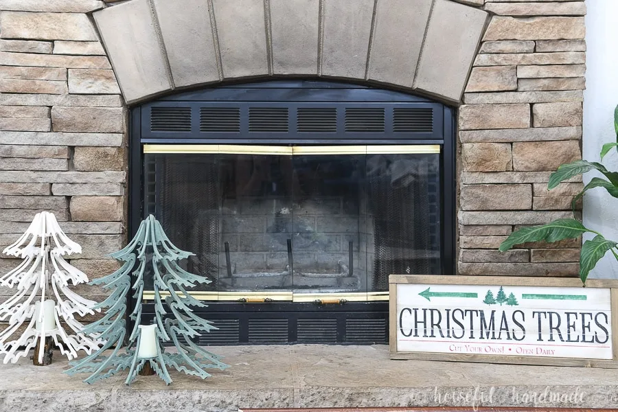 Two 3D wood Christmas tree lanterns sitting on a stone fireplace hearth with a Christmas tree farm sign on the other side of the hearth.