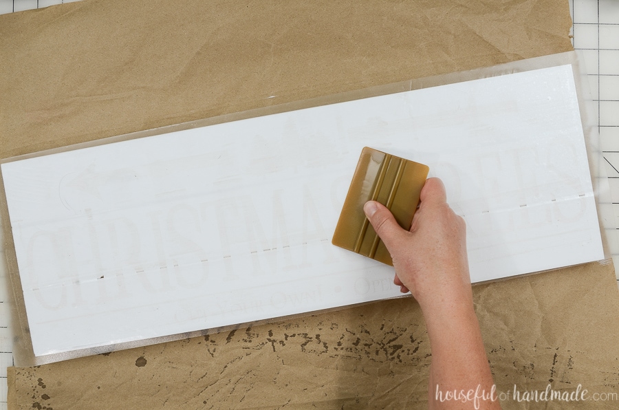 Using a vinyl scrapper to smooth the vinyl stencil over the white painted wood boards. 