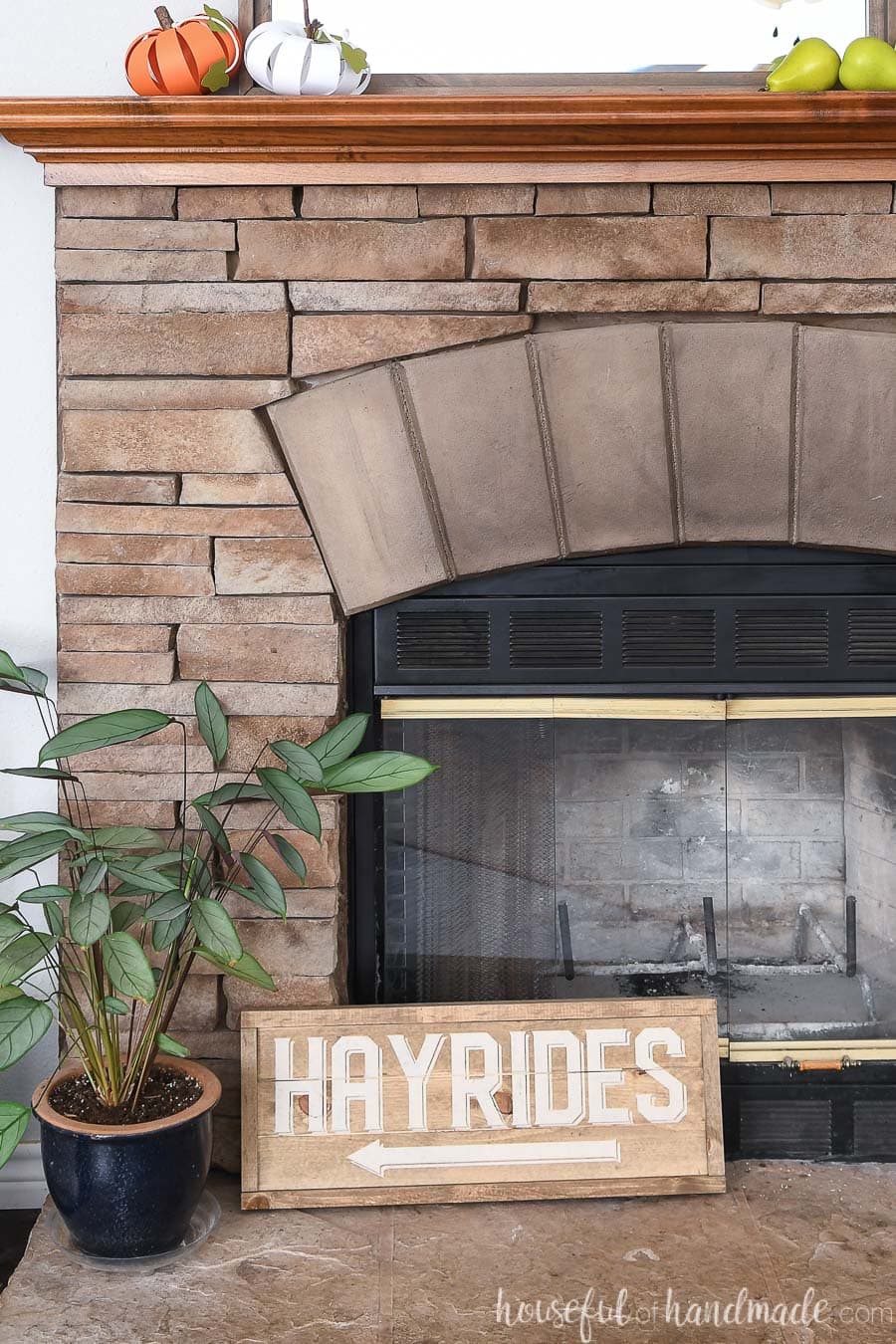 Neutral hayrides side of the double sided wood sign on a fireplace hearth next to a plant.