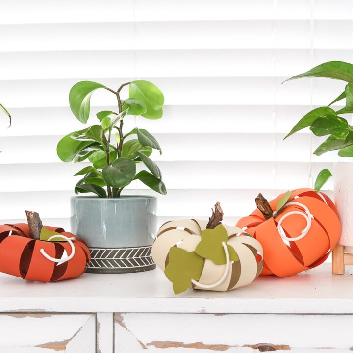 Console table with 3 paper pumpkins in oranges and cream next to two potted house plants.