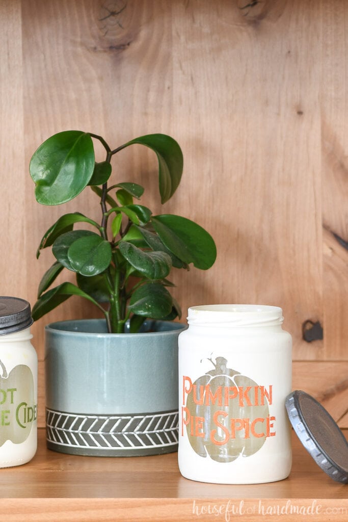 White jar with see-through pumpkin shape and orange text Pumpkin Spice Spice over it with plant and second jar in the background.