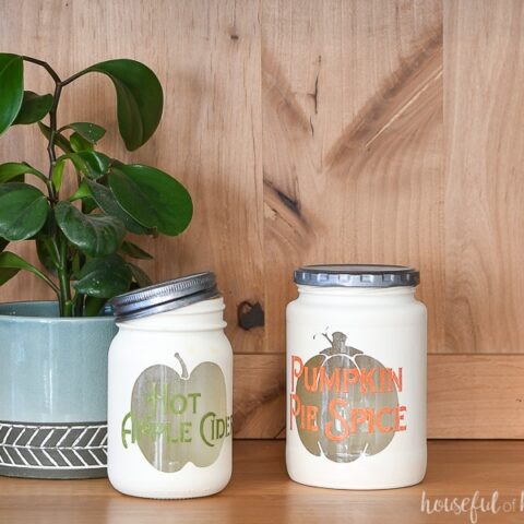 Two fall themed kitchen canisters in a wood hutch with a houseplant in the background.
