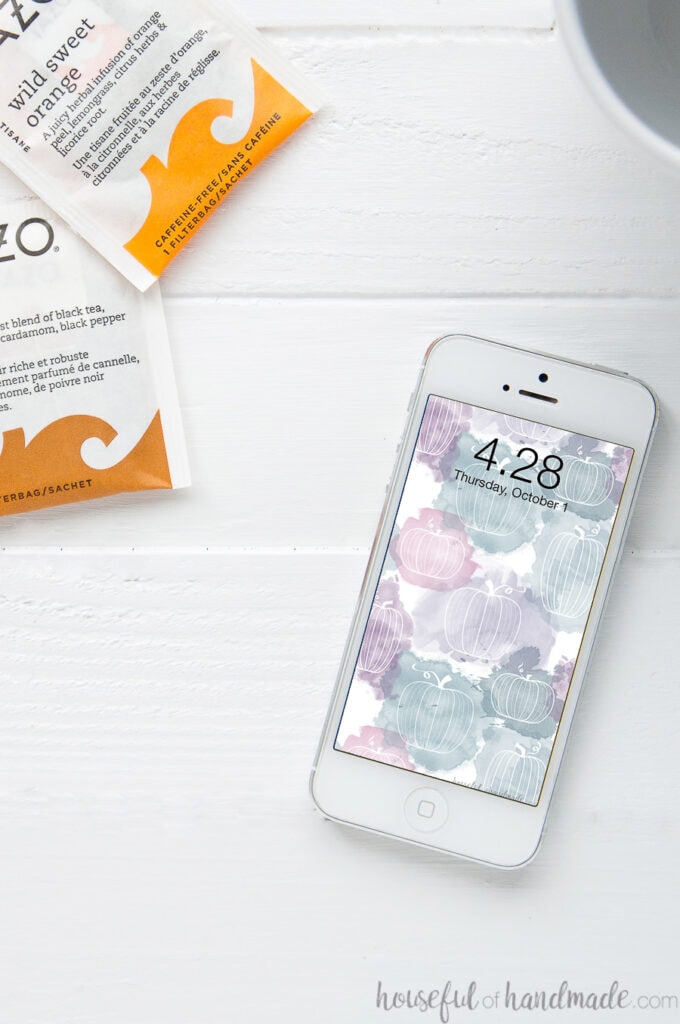 White iPhone on a white table next to fall tea pouches with the free digital wallpaper for October on the home screen.