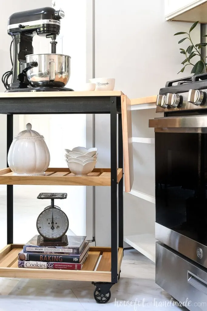 Natural wood and black rolling ktichen cart with metal corners and black wheels in a kitchen. 