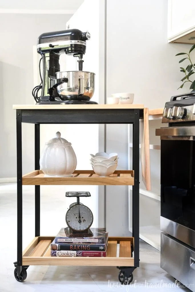 Black and wood modern rolling kitchen cart with stand mixer on top next to a stove.