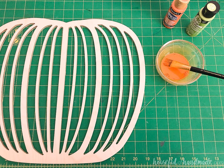Modern pumpkin design cut out of wood on a green cutting mat next to craft paint. 