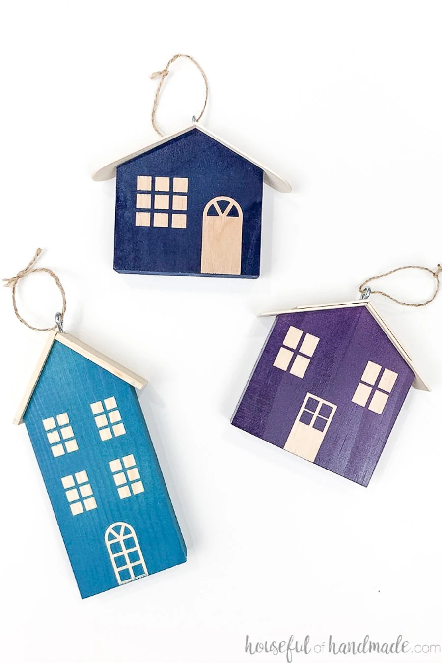 Three wood Christmas houses with eye hooks on the top to hang as ornaments, one turquoise, one navy and one purple, laying on a white background.