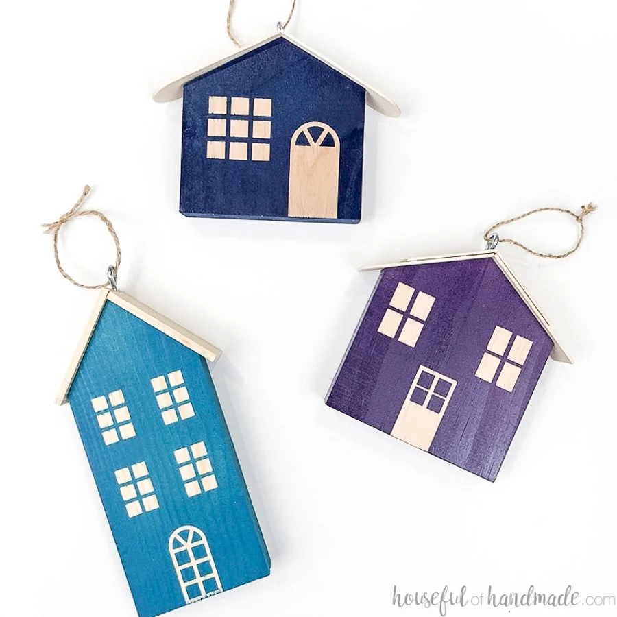 Square photo of 3 colorful wood Christmas house ornaments laying on a flat white surface. 