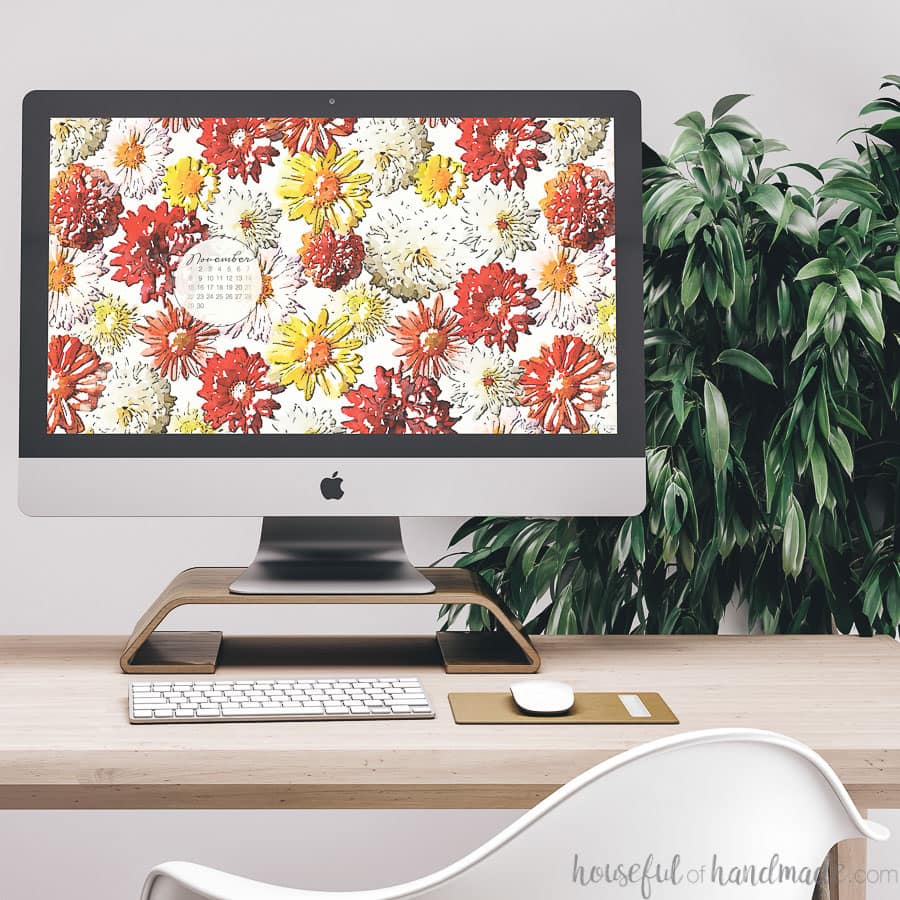 Desktop computer showing the free digital background for November on the screen sitting on a desk in front of a plant.