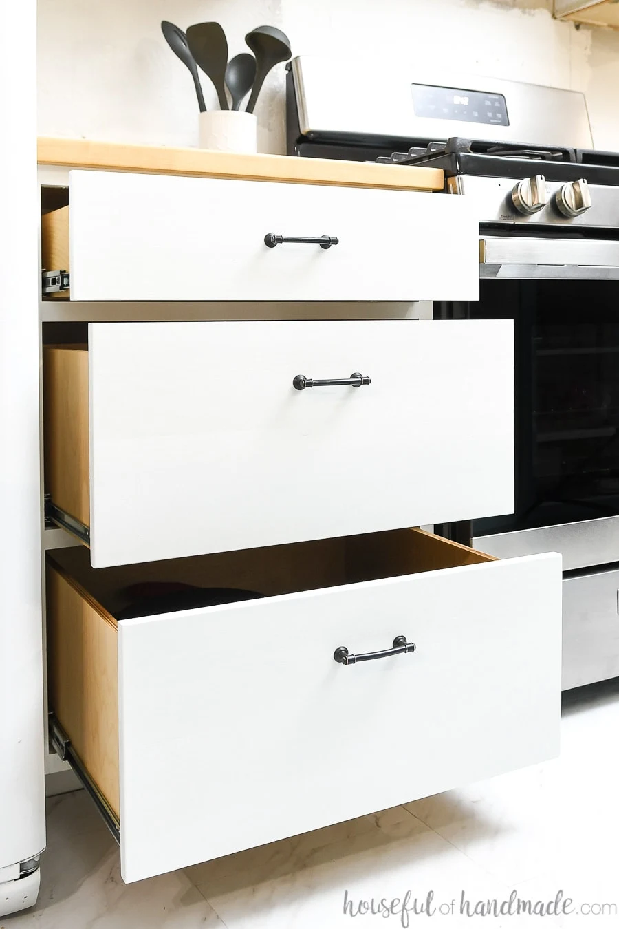 Three white kitchen cabinet drawers with black pulls pulled out partially showing the ball bearing drawer slides on the sides of the drawers. 