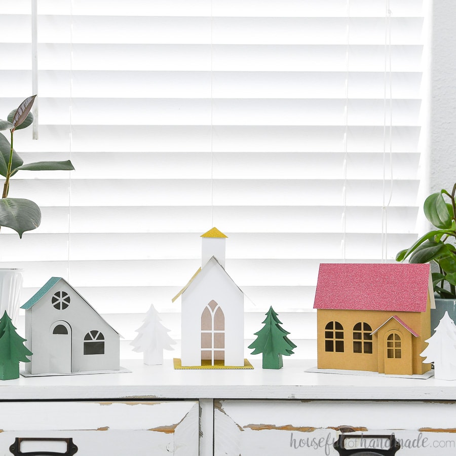 Putz inspired paper Christmas village with 3 different houses and paper Christmas trees on a console table.