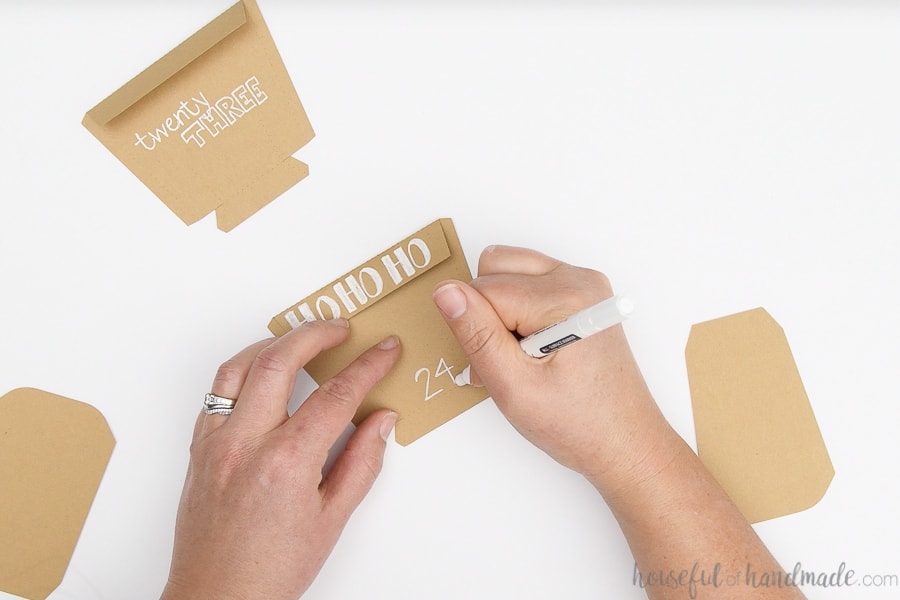 Paper pieces cut out of brown Kraft cardstock being decorated with a white paint pen with advent calendar numbers and Christmas pictures.