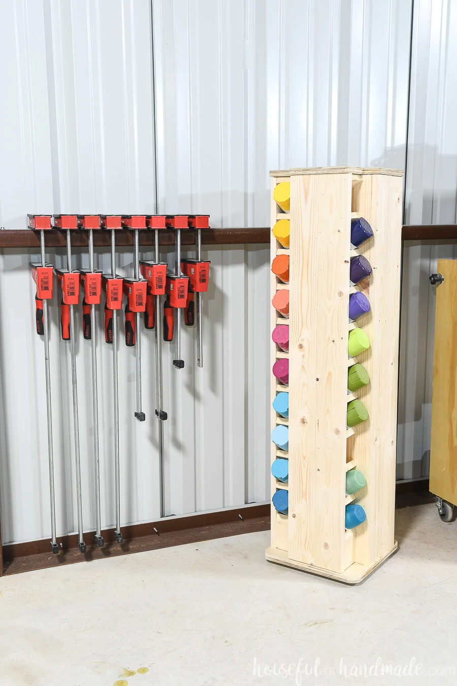 Small rotating spray paint storage cabinet with spray paint cans in organized in a rainbow in a corner next to bar clamps in a shop.