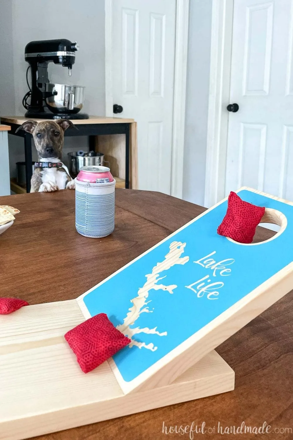 Close up of the one side of the miniature cornhole board on a table with red bean bags on the board and a puppy looking at it in the background.