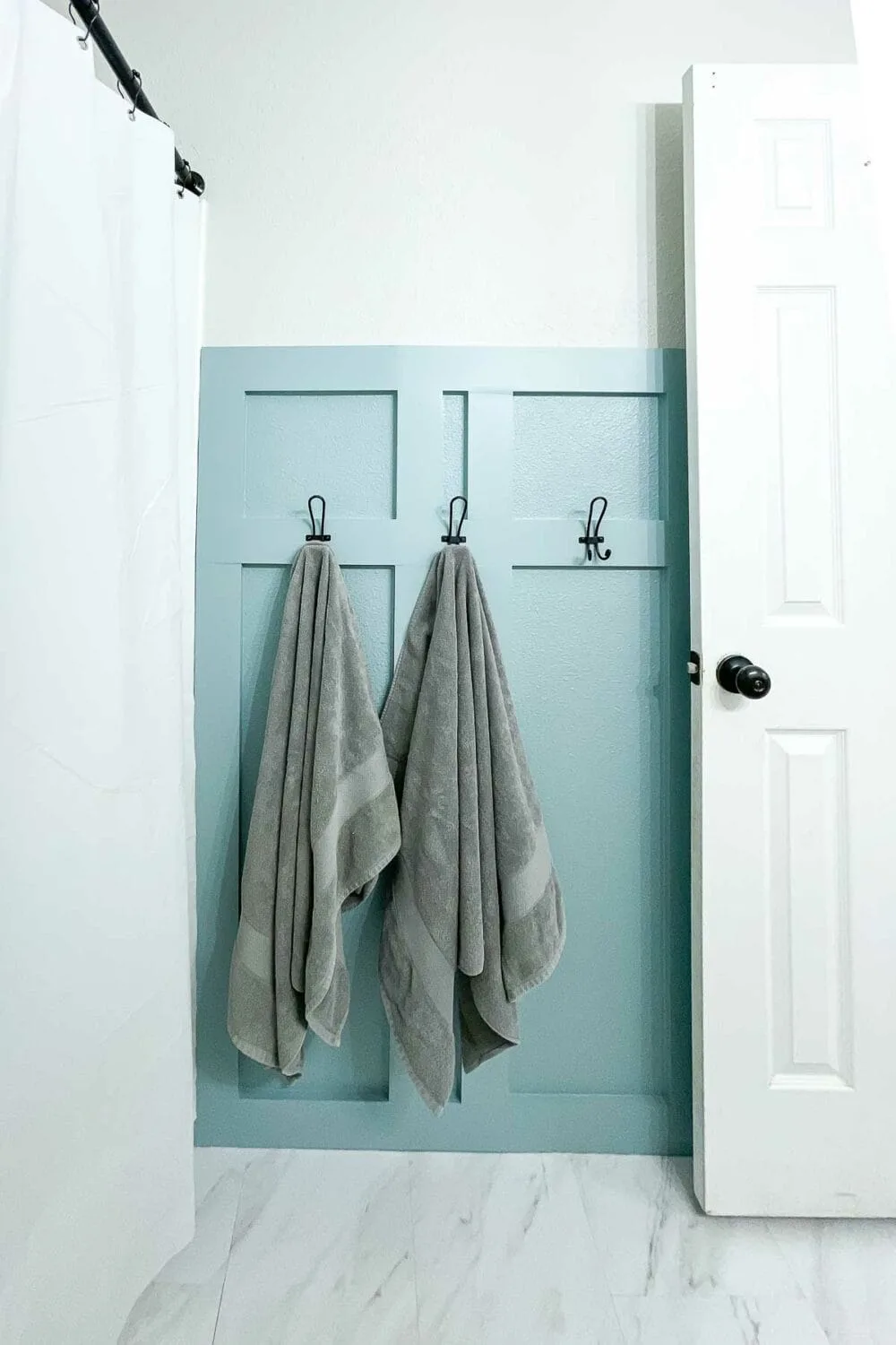 Blue painted board and batten wall in the bathroom with black hooks on it and gray towels hanging on 2 of the hooks.