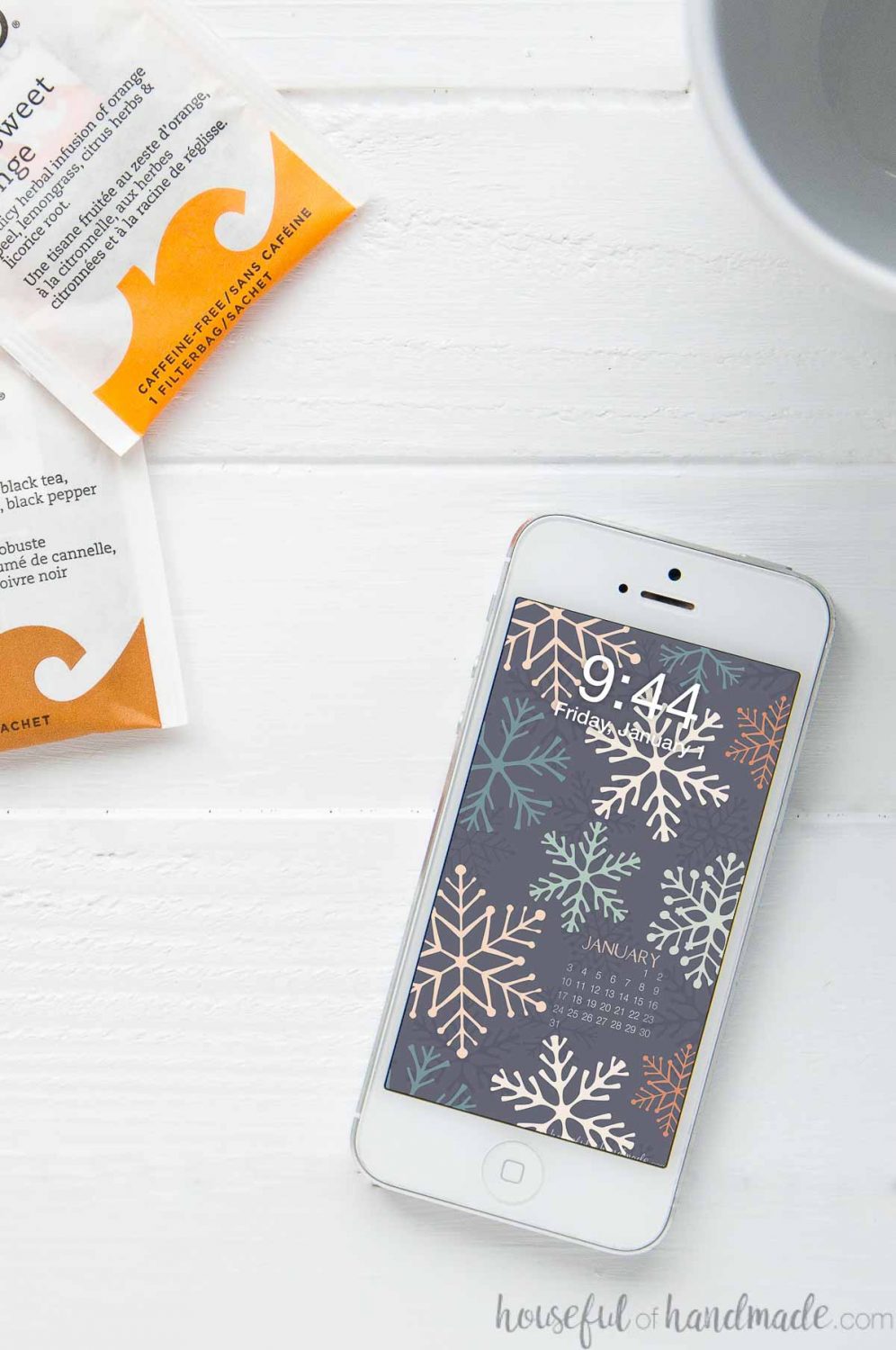 White smartphone with the free digital wallpaper for January with a calendar on the screen laying on a table by tea packets and a mug.
