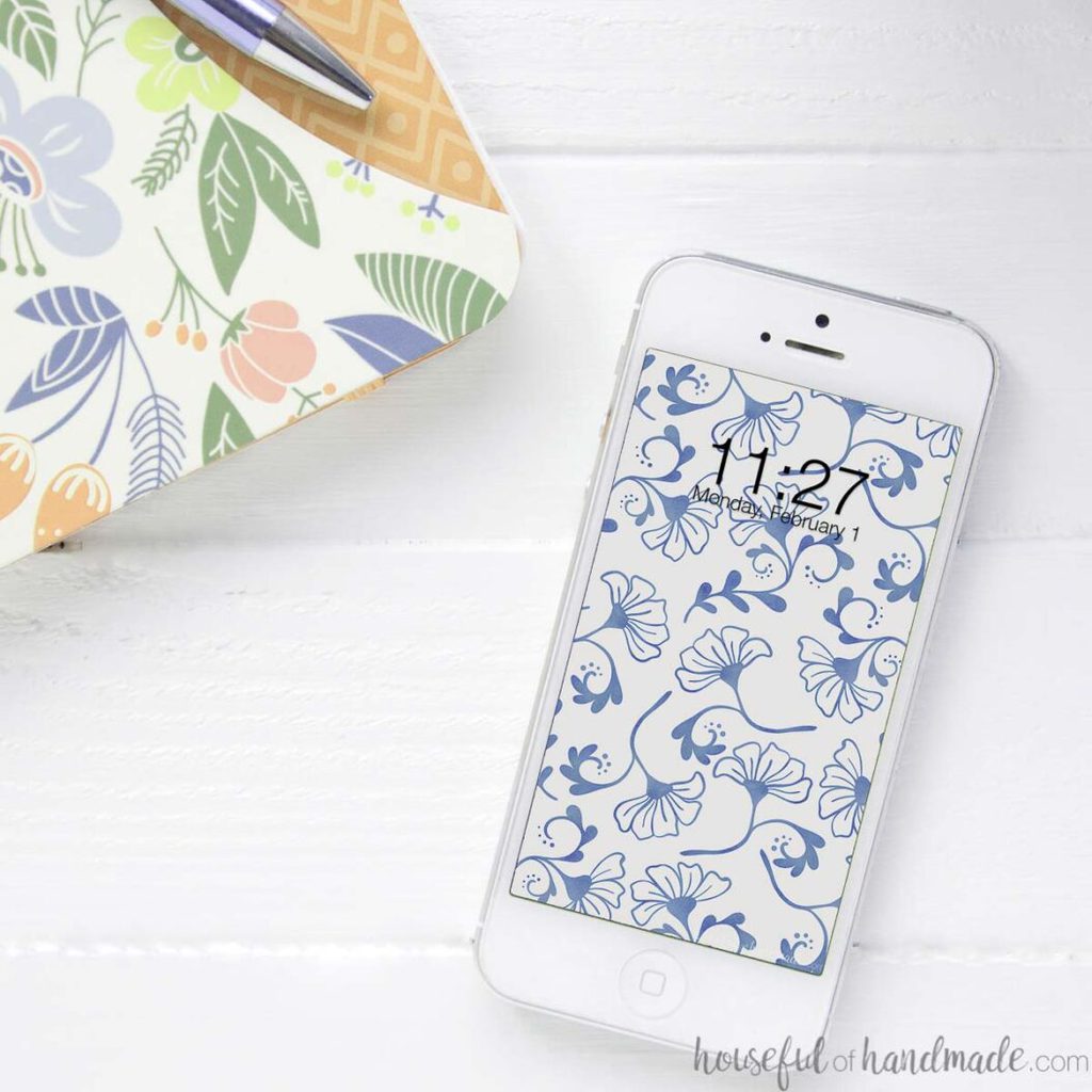 Smartphone with the blue floral digital background on the home screen on a desk next to a notebook. 