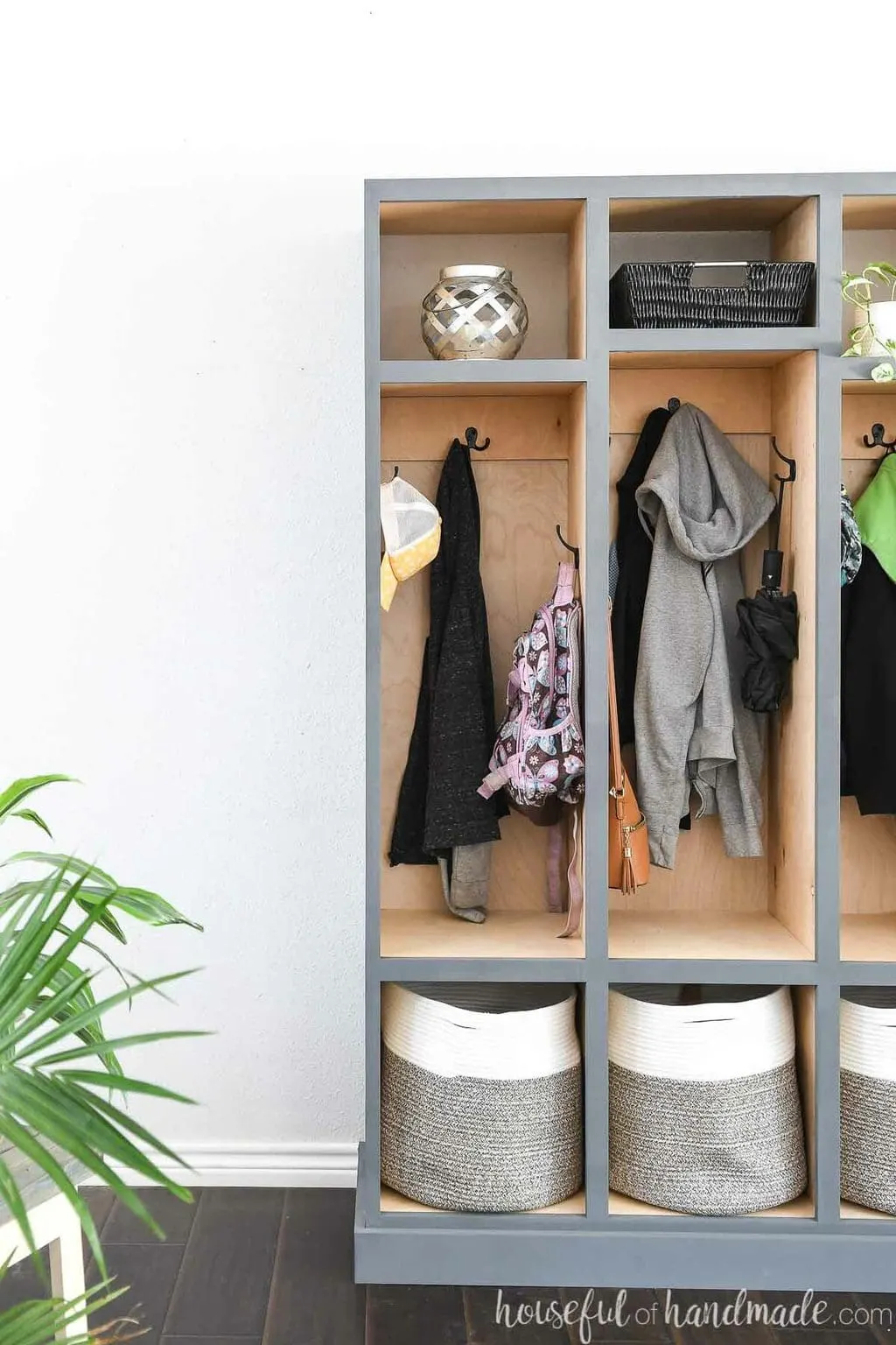 Looking at just half of the mudroom storage lockers with sweaters and backpacks hanging on the hooks and a fern in the foreground.