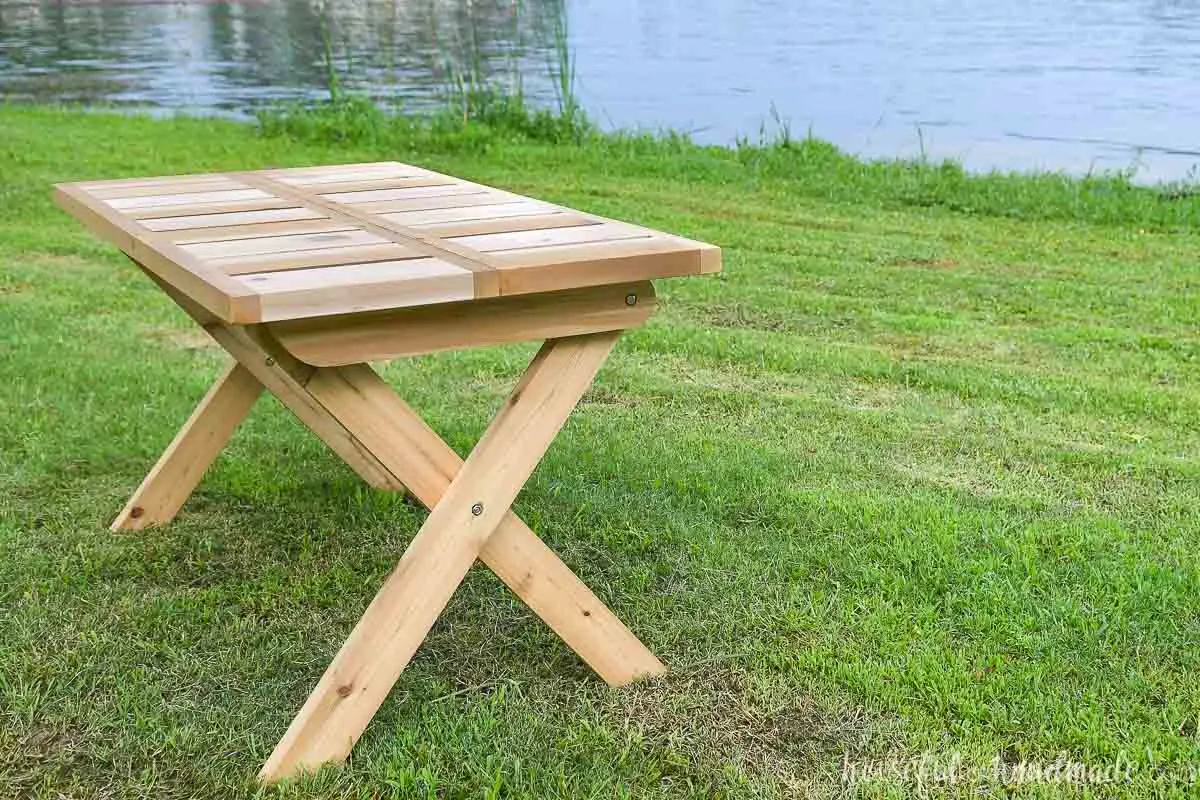 X-leg folding picnic table made from wood sitting on the lawn by a lake. 
