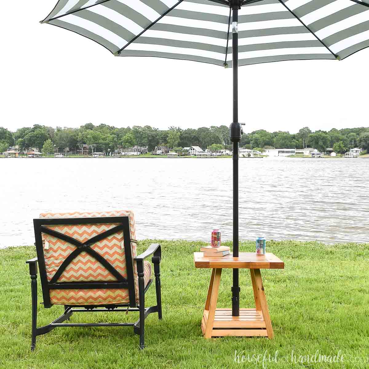 DIY umbrella side table with black and white striped umbrella in it and outdoor chair with cushions next to it looking over a lake.