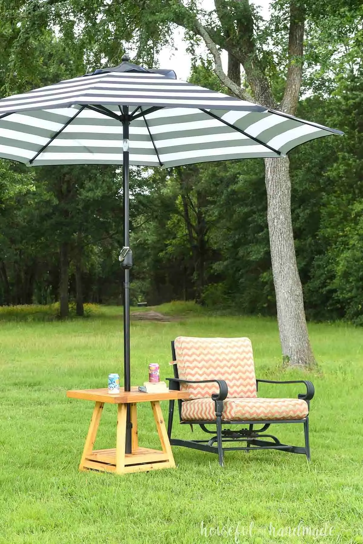 Small outdoor umbrella table next to chair on a lawn with trees in the background. 