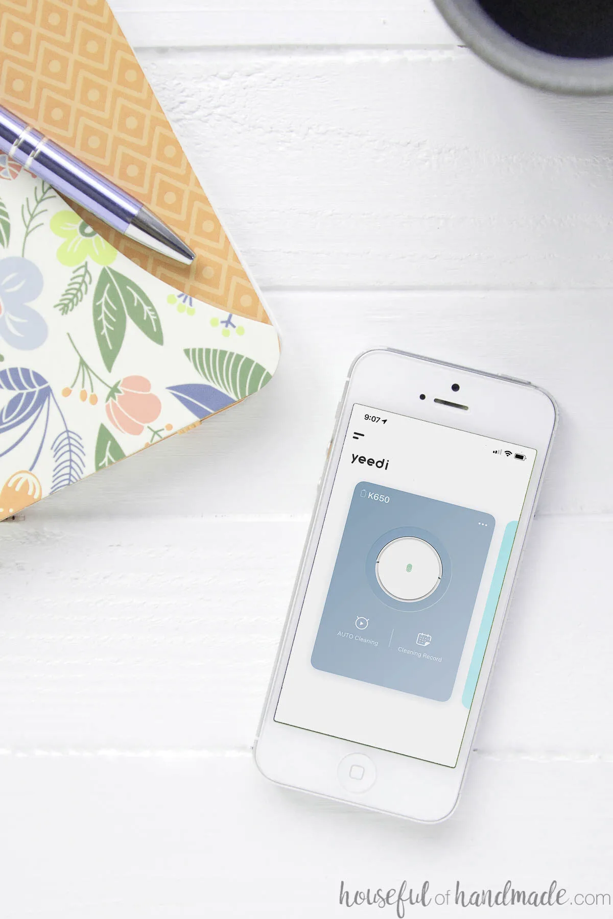 White cell phone showing the Yeedi Robot vacuum app on the screen next to a notebook and coffee mug.