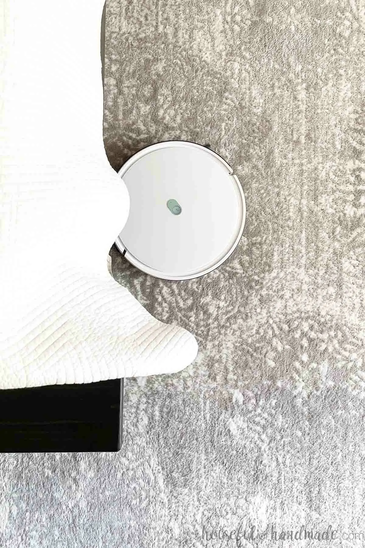 Top down view of a white robot vacuum cleaning a rug under a bed. 