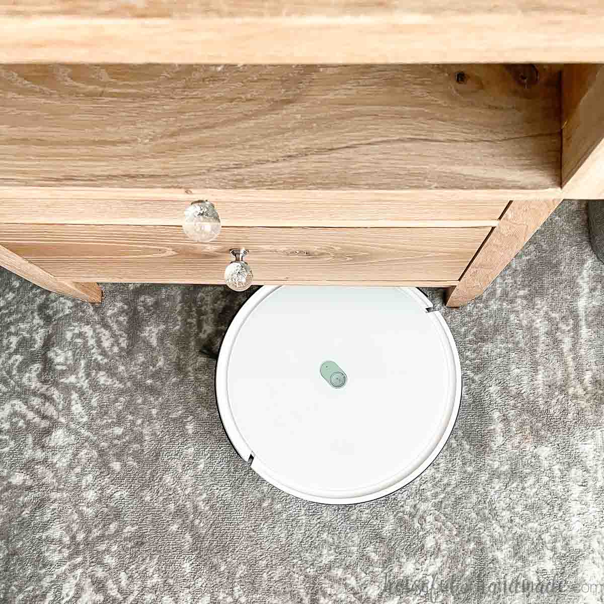 Looking down at the front of a nightstand with a white Yeedi robot vacuum cleaning the gray and white rug below it.