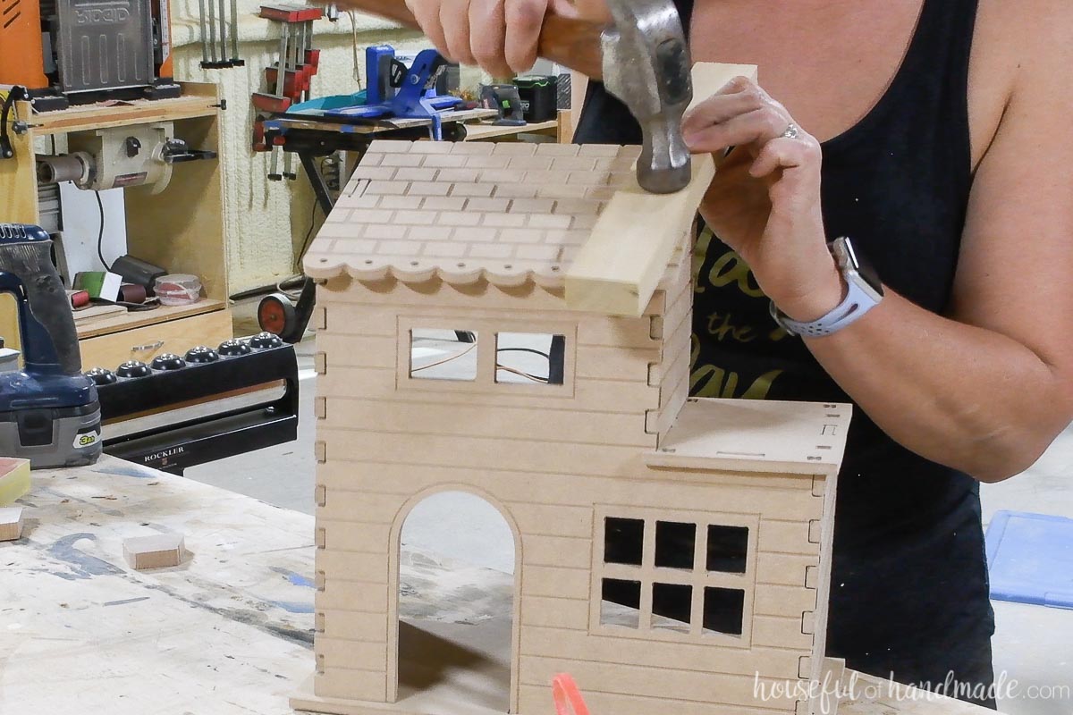 Using a hammer and scrap of wood to tap the tabs into the notches on the roof of the MDF doll house. 