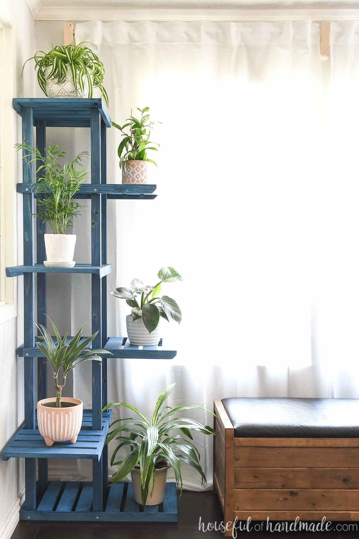 Tall corner plant stand full of indoor house plants in a corner next to a bench in front of a window. 