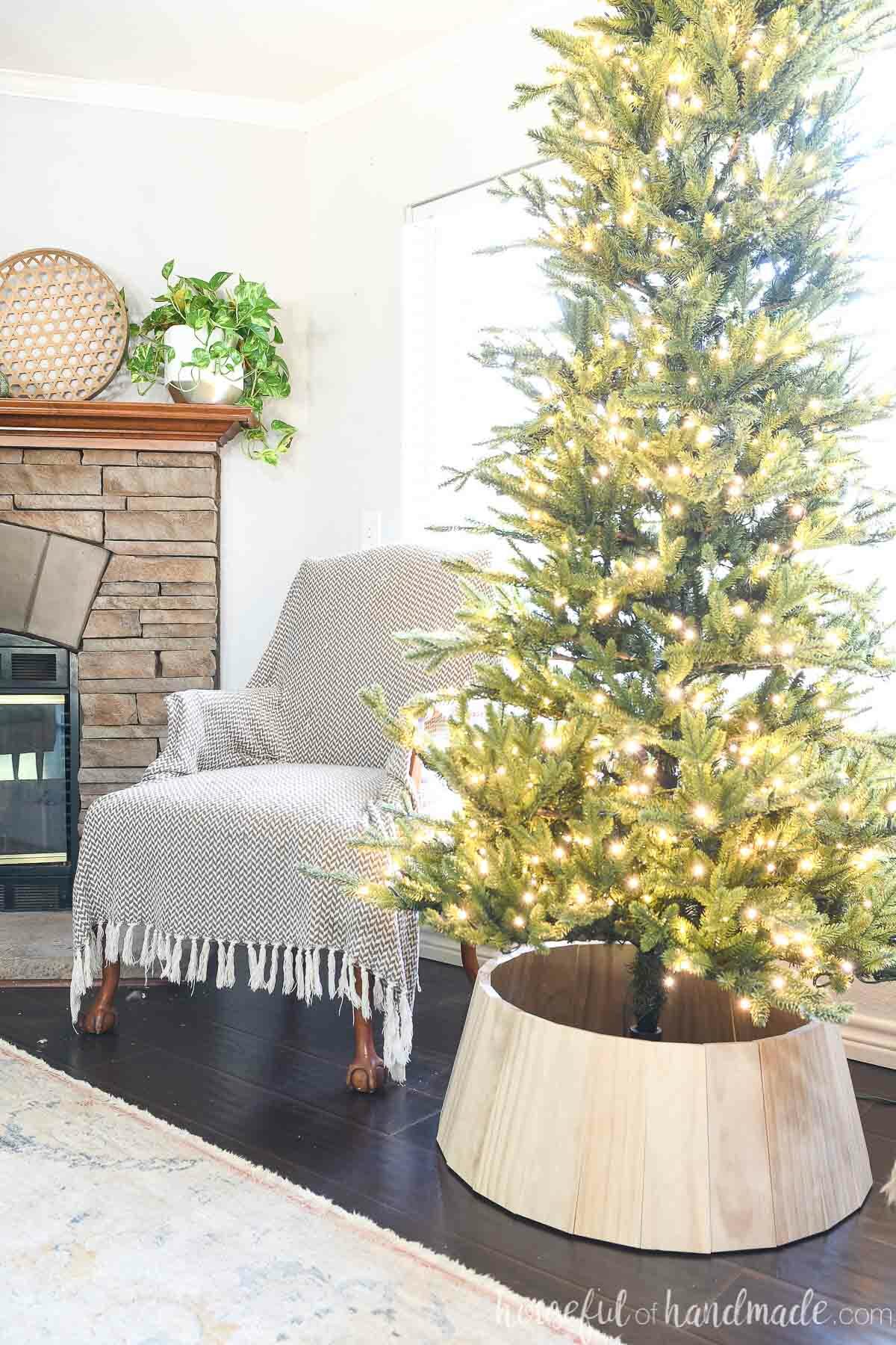 Living room with a Christmas tree with a wood tree collar and chair next to a fireplace. 