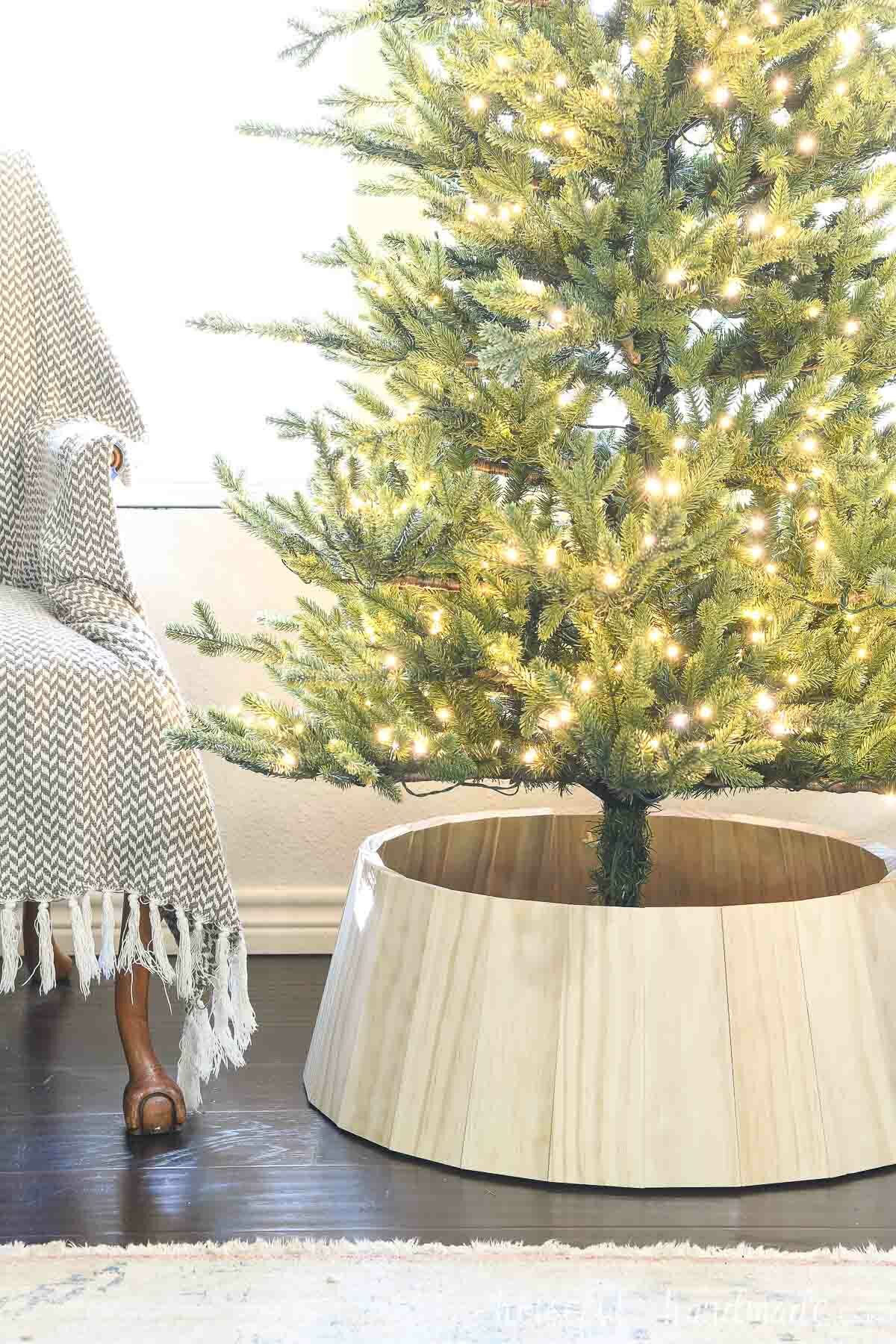 DIY wooden Christmas tree collar around the base of a slim tree next to a chair. 