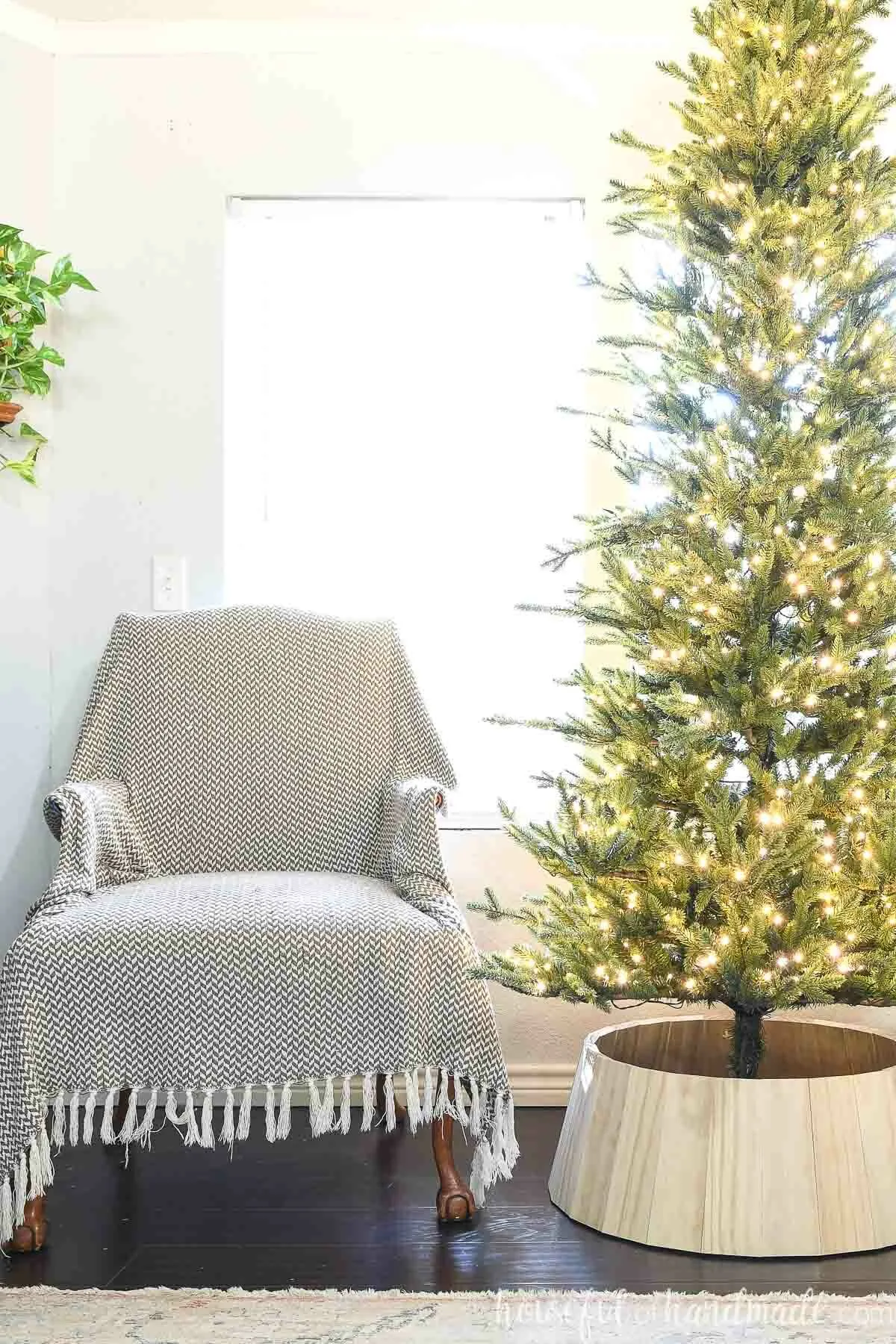 Slim pre-lit faux Christmas tree with wood tree collar next to a chair. 