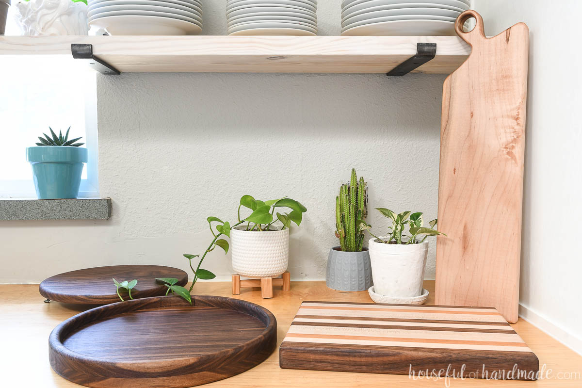 Three easy handmade woodworking gifts sitting on a countertop in a kitchen with open shelves above and plants around them. 