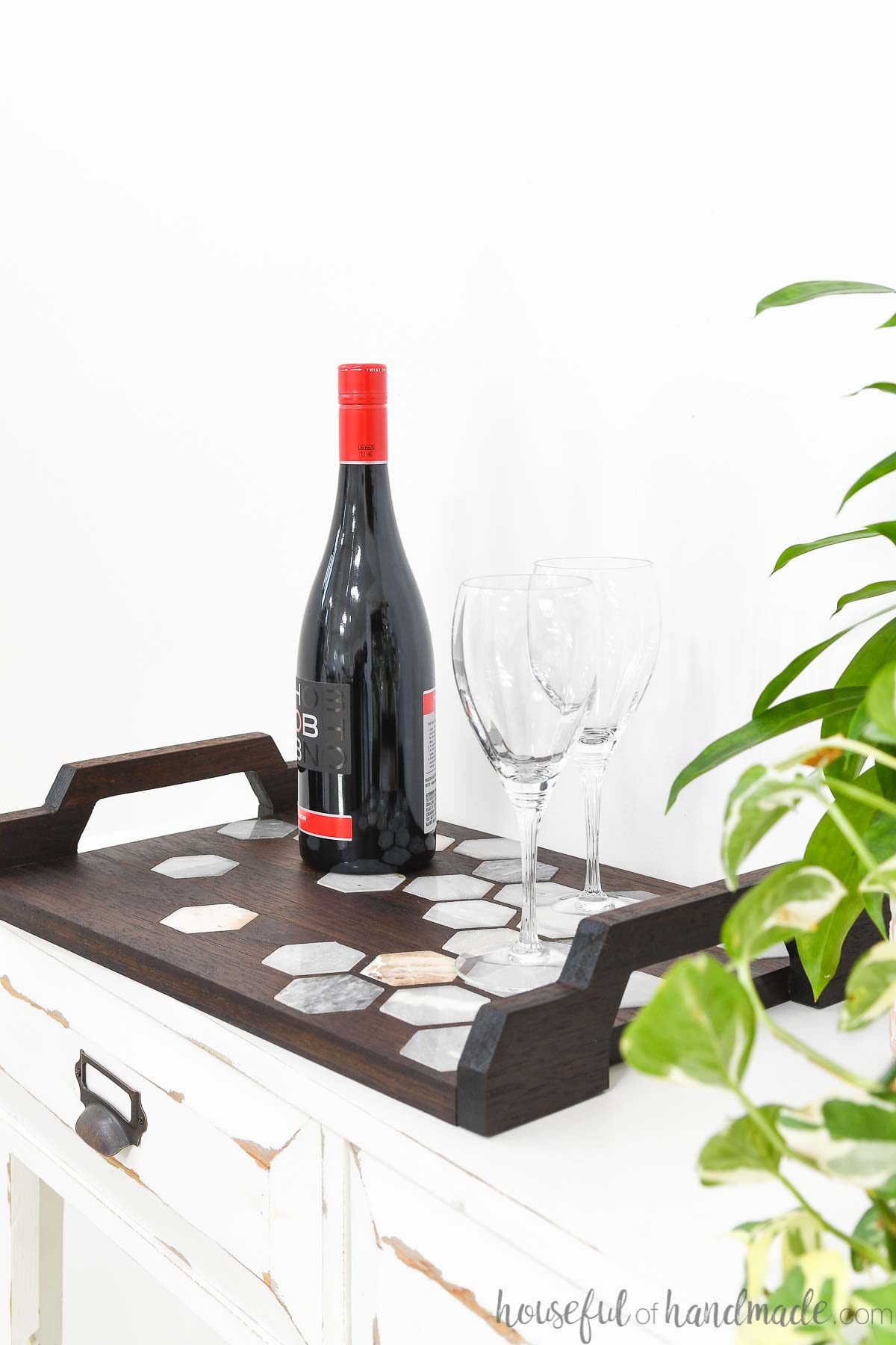 White side table holding a wood and marble tray with wine on it next to house plants. 