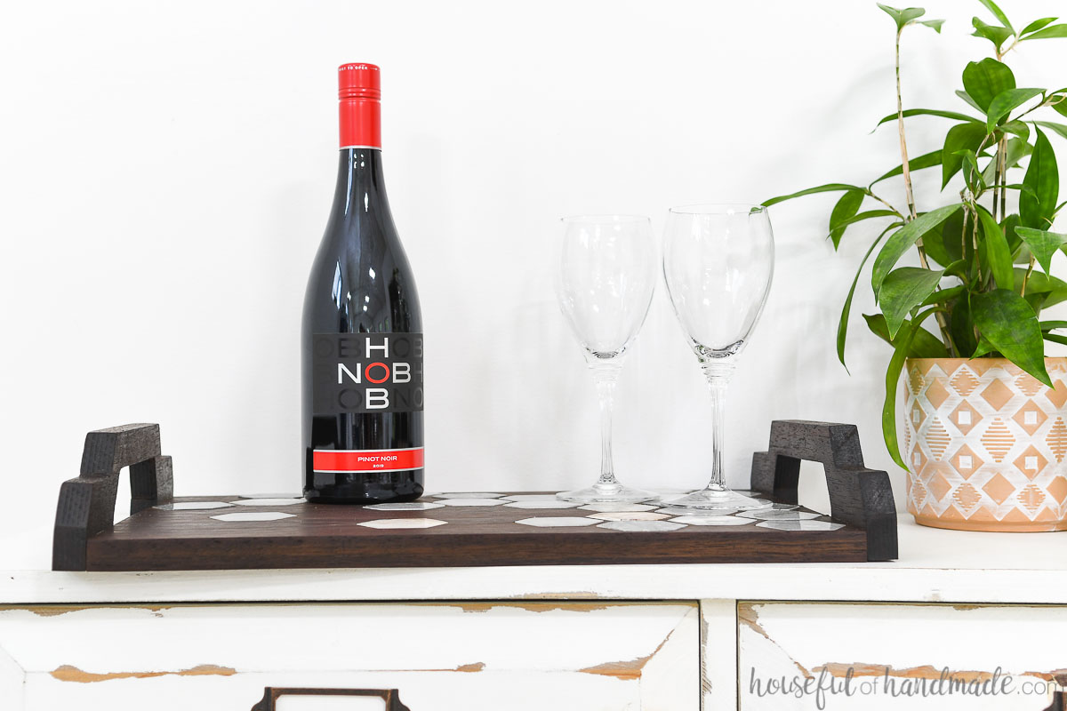 DIY wood serving tray with handles holding white glasses and a bottle of wine on a side board next to a plant. 