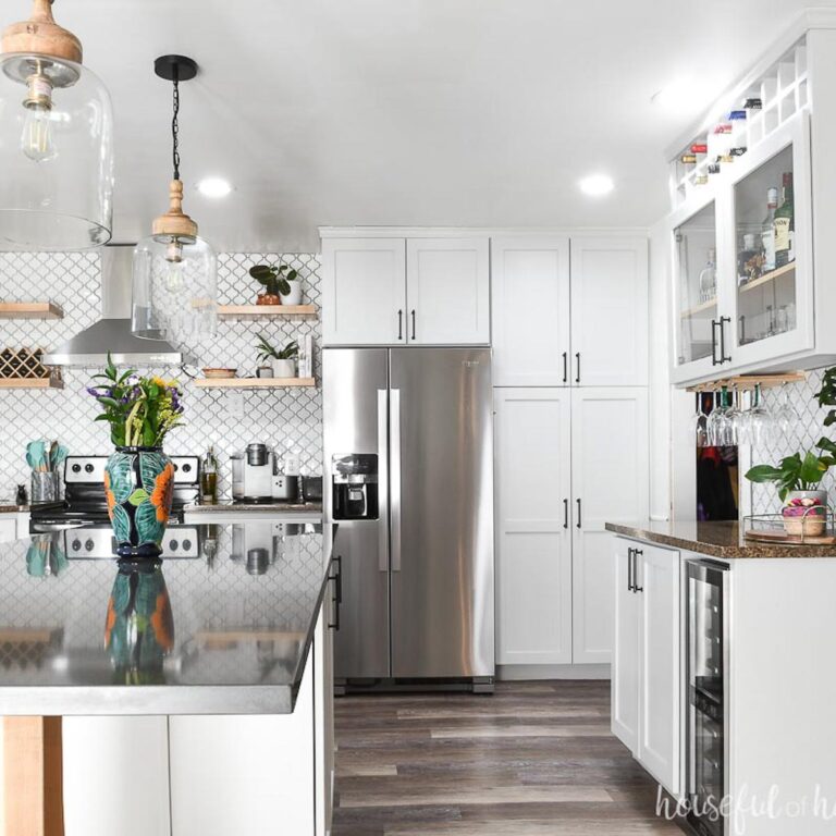 Modern kitchen with pale gray painted cabinets, white arabesque tile wall, vinyl plank flooring and dark solid surface countertops.