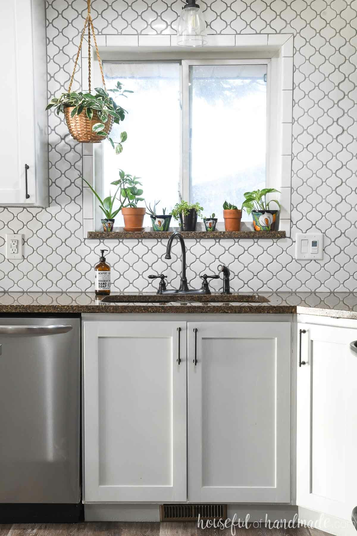 Pale gray kitchen sink cabinet blow a window with white porcelain tile filling the wall to the ceiling and framed around the window. 