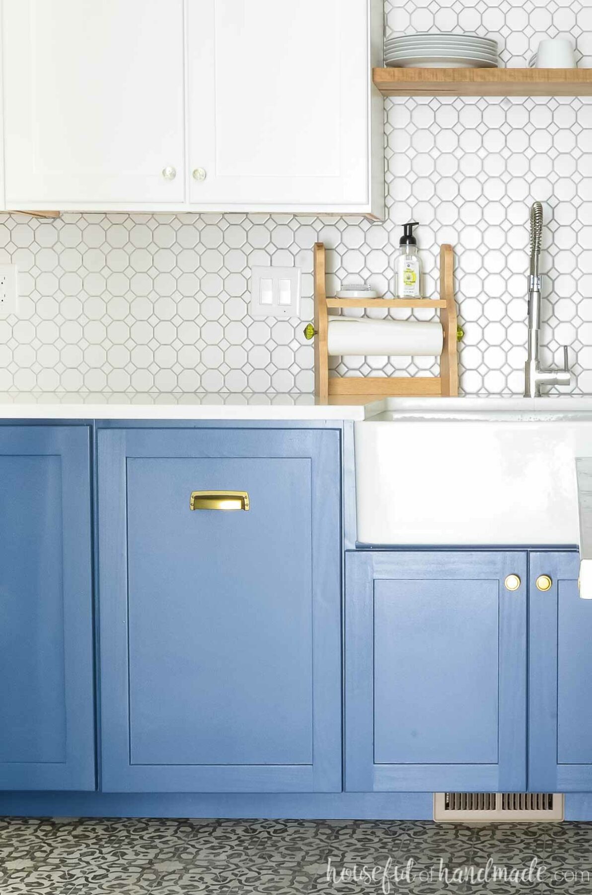 Blue and white kitchen with trash cans hidden behind a cabinet door next to a white farmhouse sink.