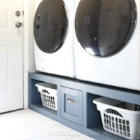 Navy blue washer dryer platform with a drawer in the center of two shelves to hold laundry baskets.