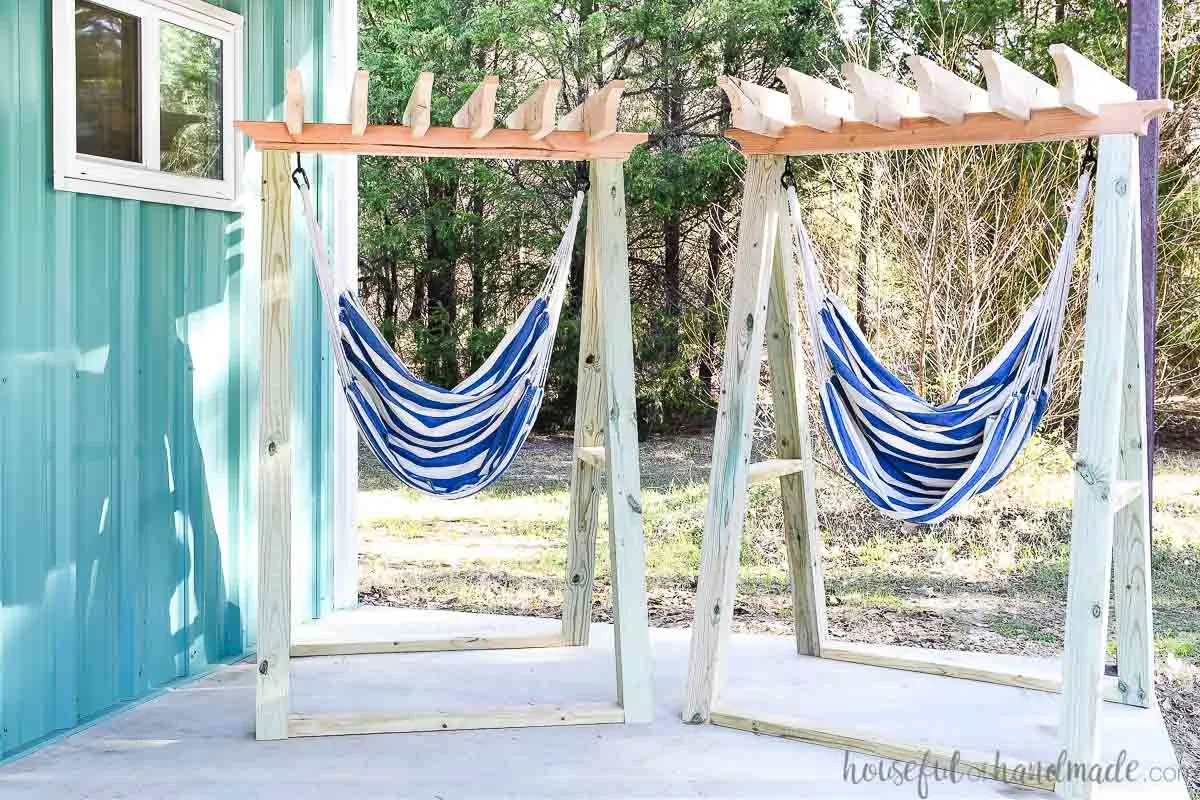 Two free standing hammock chair stands sitting on a porch in front of an aqua metal building. 